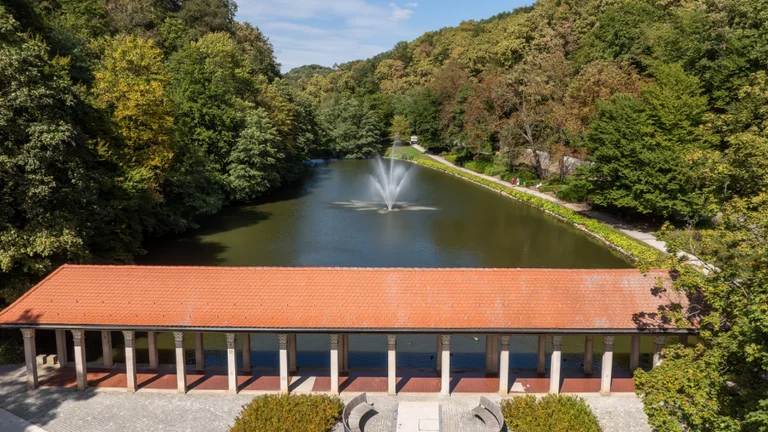 Maribor city park featuring a pond with a fountain, surrounded by lush trees and a charming pavilion with a red-tiled roof.
