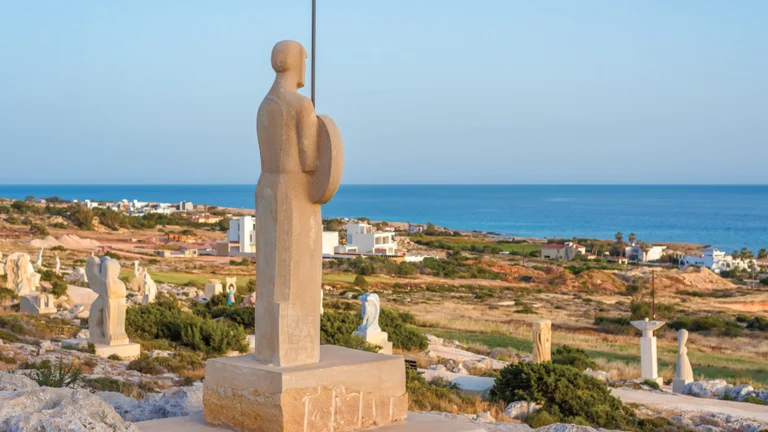 Kavo Gkreko Sculpture Park., overlooking a coastal landscape with scattered sculptures and homes in the distance.