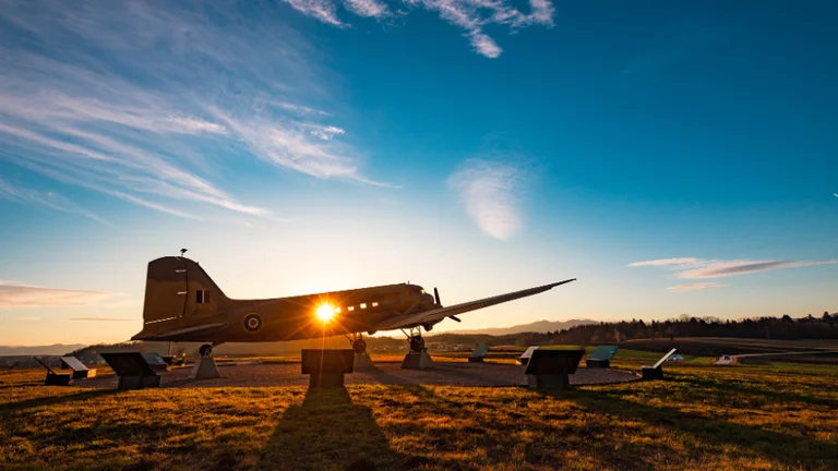A vintage aircraft silhouetted against a sunset, surrounded by informational plaques on a grassy landscape.