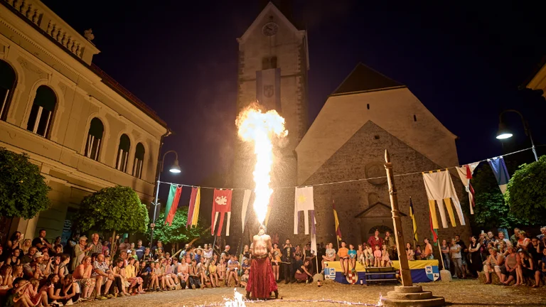 A fire performer breathes flames in a bustling square, surrounded by an engaged crowd and illuminated by the night sky.