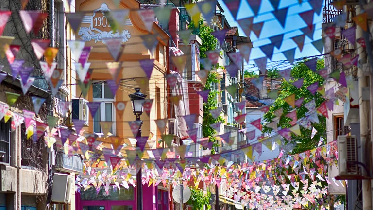 A festive street view in Kapana, Plovdiv, with vibrant triangular flags strung overhead and colourful historic buildings in the background.