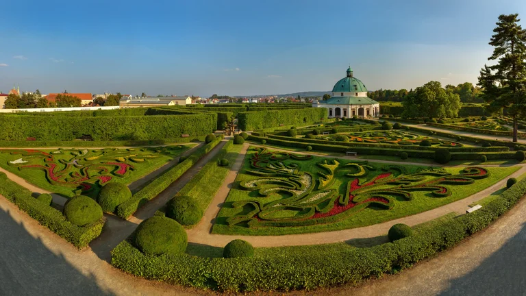 A panoramic view of a beautifully manicured garden with vibrant floral designs and a dome-building in the background under a blue sky.