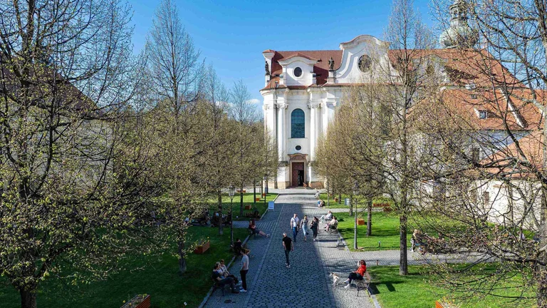 A sunny plaza with people walking and sitting on benches, framed by newly leafed trees and the baroque façade of a church in the background.