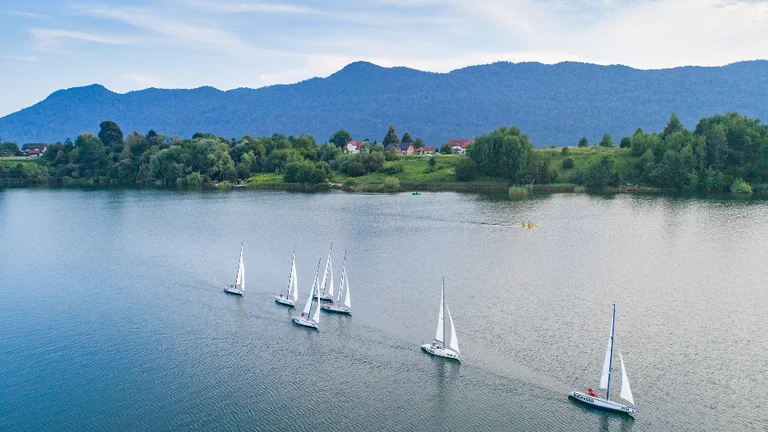 Sailboats gliding on the calm waters of Kočevsko Lake, Slovenia, surrounded by lush forest and distant hills.