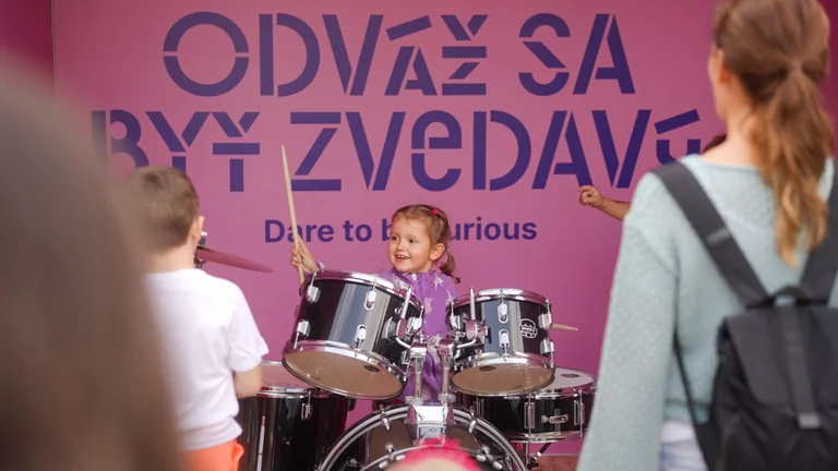 A young girl enthusiastically playing a drum set in front of a pink sign that reads "Dare to be Curious" at an outdoor event.