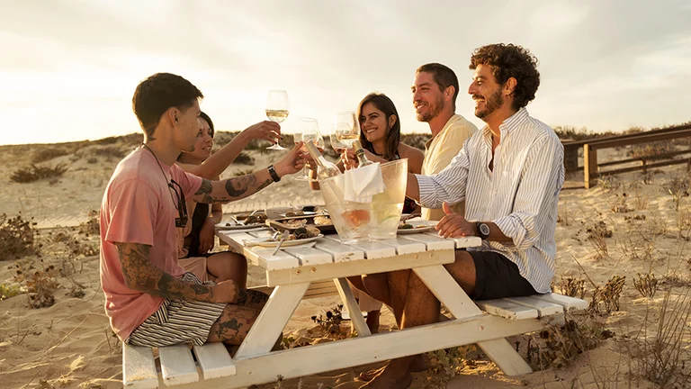 A group of friends sitting at a wooden bench table on the beach, smiling and cheering glasses of wine during a sunny day.