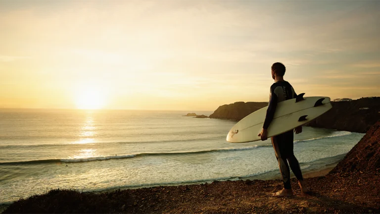 Surfer standing on the sandy beach of Praia da Arrifana, Algarve, Portugal, with cliffs in the background and no visible waves.