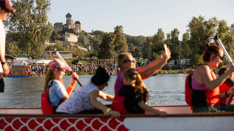 Dragon boat racing with people paddling on a river, with Trenčín Castle visible on a forested hill in the background.