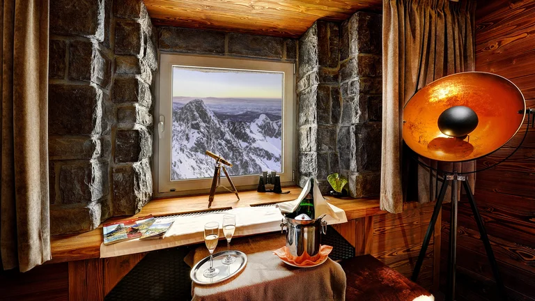 Cosy mountain lodge interior with stone walls and wooden furnishings, overlooking Lomnický štít peak in the High Tatras, Slovakia, with champagne by the window and snow-covered mountains outside.