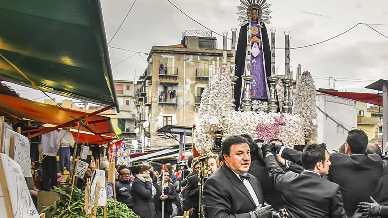 Men in suits carry a religious statue through a crowded outdoor market under cloudy skies.