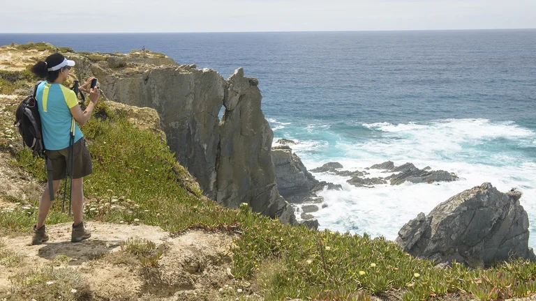 Person taking a photo on a rocky seaside cliff with waves crashing below.