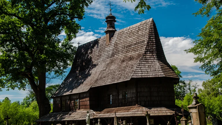 Old wooden church with steep roof surrounded by trees under blue sky.