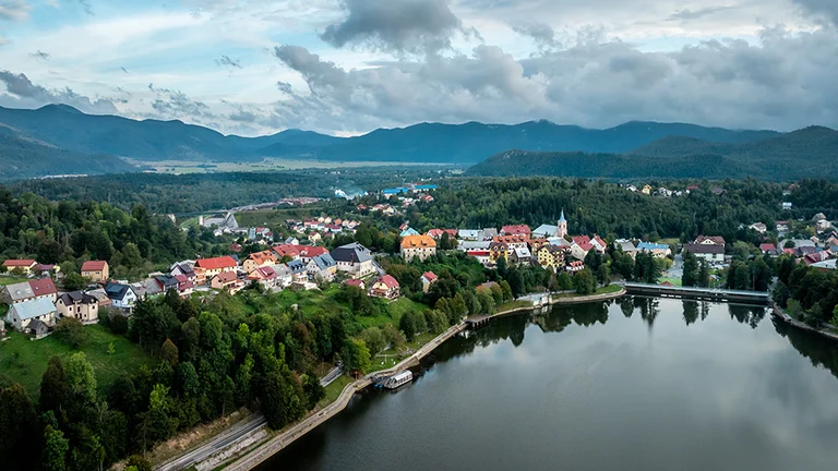 Colorful village houses by a calm lake, surrounded by forested hills under a cloudy sky.