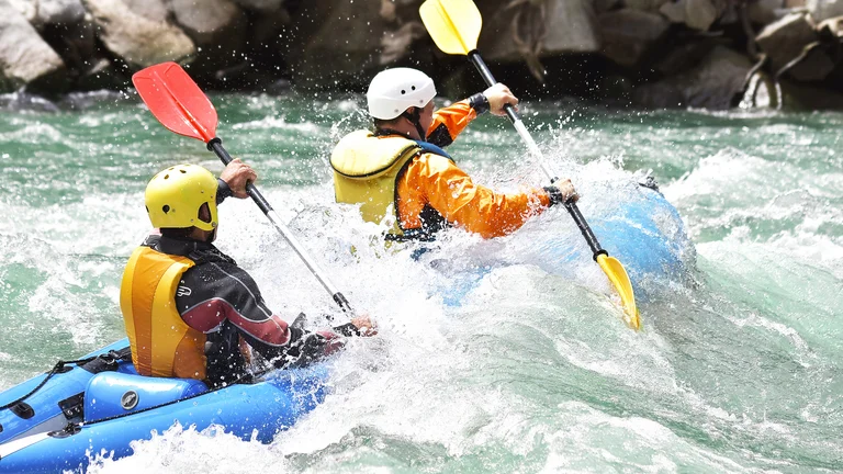 Two people kayaking through whitewater rapids, wearing helmets and life jackets.
