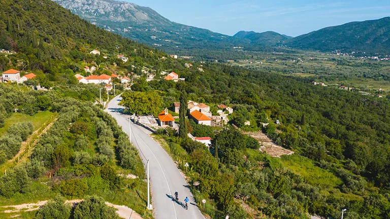 Winding road through a green valley with houses and mountains in the background.