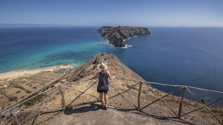 Person standing at a cliffside viewpoint overlooking blue ocean and rocky island.