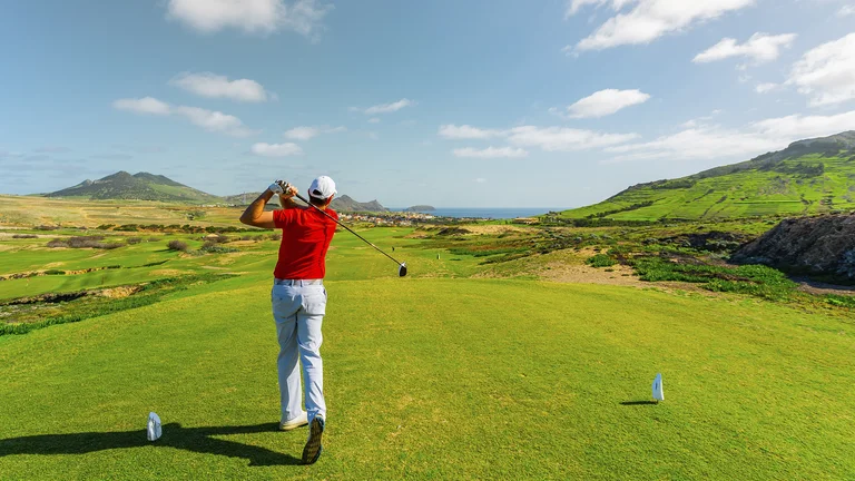 Golfer in red shirt swings on a sunny, green golf course with rolling hills.