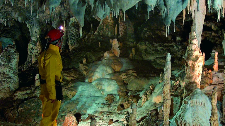 Person in yellow suit exploring a cave filled with stalactites and stalagmites.