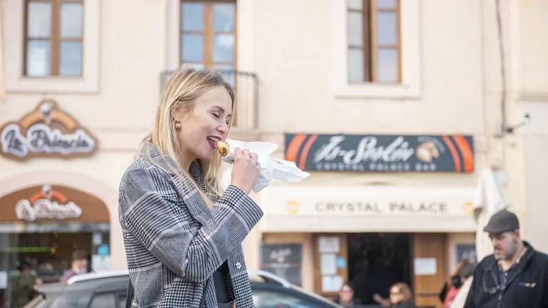 Woman in a gray plaid coat eating a sandwich on a city street.