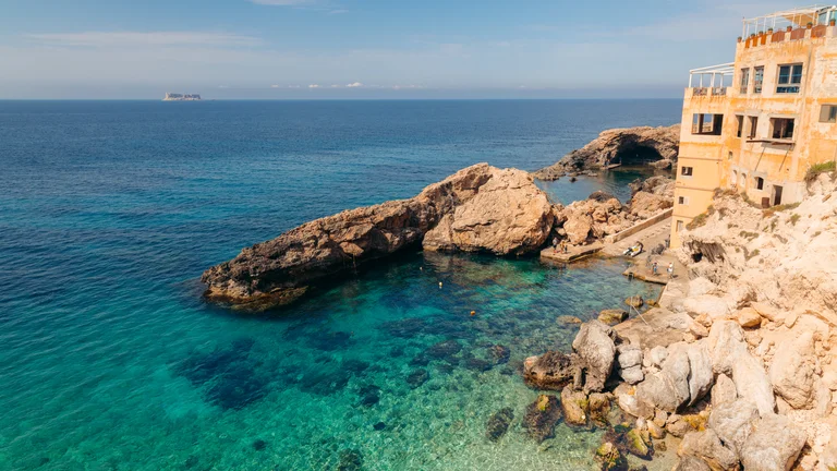 Clear blue sea with rocky coastline and a tan building on the right under a sunny sky.