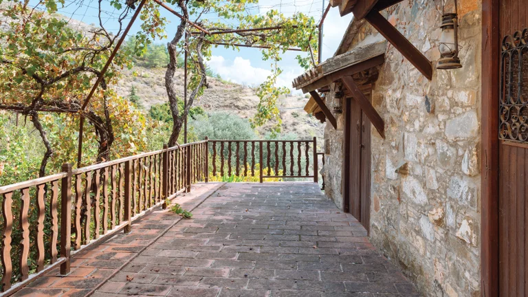 Rustic stone balcony with wooden railing and leafy vines, overlooking a sunny hillside.