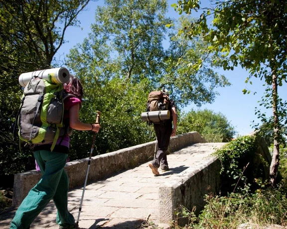 Two hikers with large backpacks trekking across an old stone bridge surrounded by lush greenery and trees in what appears to be a serene natural environment, suggesting an outdoor adventure on a bright sunny day.