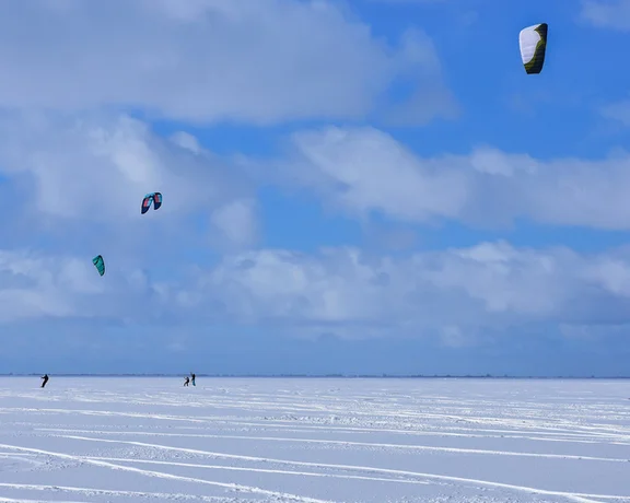 People snowkiting on a frozen lake with colorful kites against a blue sky.