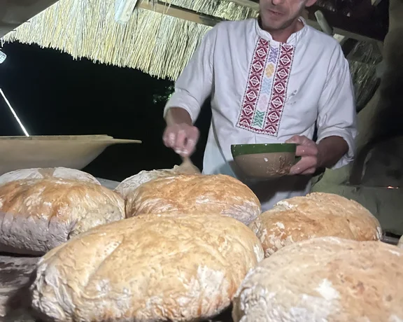 A person in traditional attire prepares bread on a wooden table under a thatched roof at night, with freshly baked loaves in view.