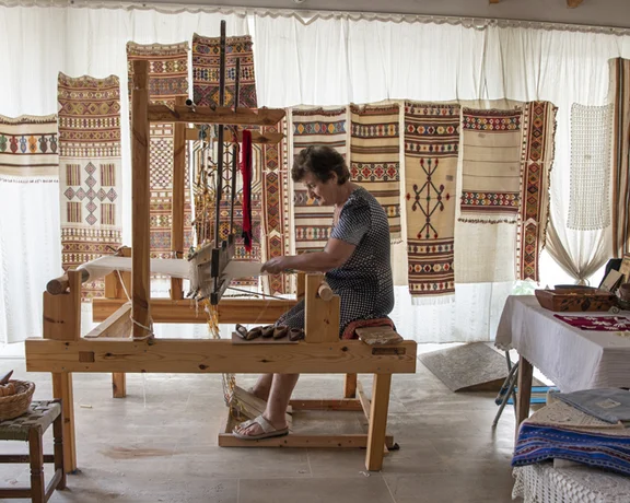 A textile artisan works at a wooden loom, surrounded by colorful woven fabrics and craft materials in a bright, airy workshop.