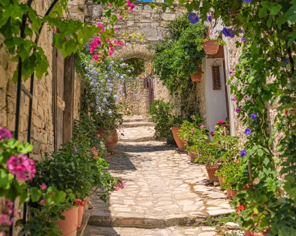 A charming cobblestone alley adorned with vibrant flowers and lush greenery, leading to a sunlit archway in a quaint village.