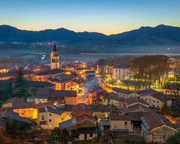 Twilight view of a tranquil village nestled amid mountains, featuring illuminated rooftops and a church steeple.