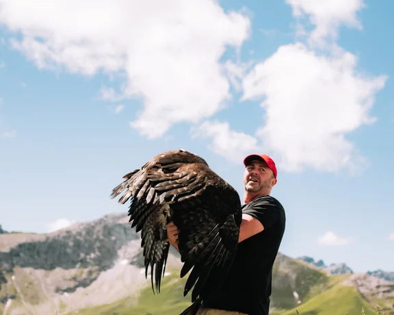 A man wearing a red cap stands in a mountainous landscape, holding a eagle with outstretched wings against a blue sky.