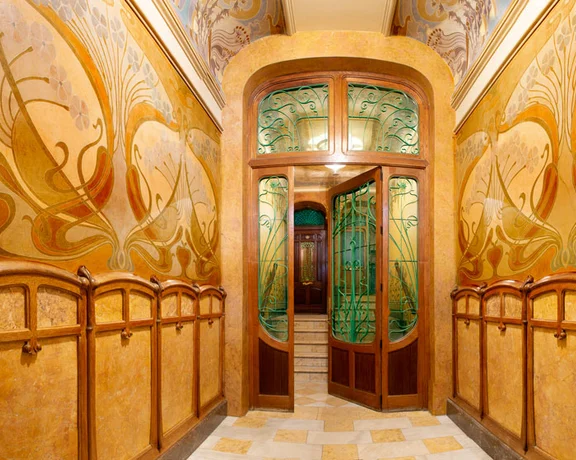 Art Nouveau hallway featuring ornate painted walls, wooden paneling, and an elegant glass door, exuding historic charm and artistry.