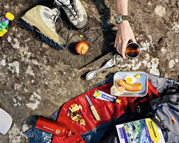 A hiker’s picnic spread on a rocky surface with hiking boots, snacks, a thermos, and trail map, capturing an outdoor meal in the Swiss Alps.