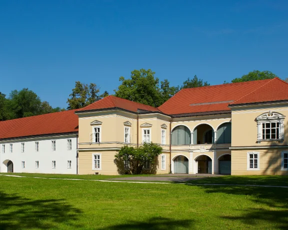 A historic building featuring a red-tiled roof, arched balconies, and lush greenery, set against a clear blue sky.