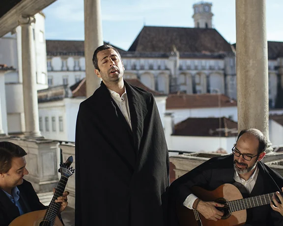 Three musicians on a balcony perform with a backdrop of historical architecture and a clear blue sky.