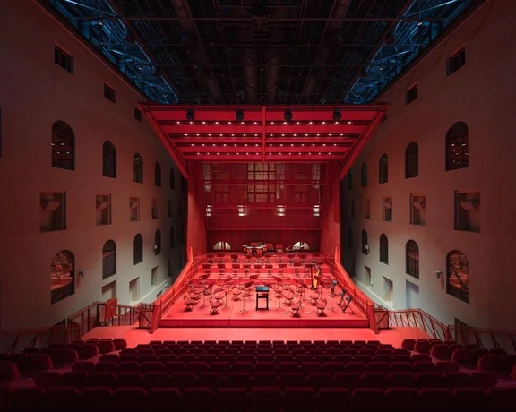 An illuminated concert hall with red hues, featuring empty chairs on stage and rows of red seats in the audience area.