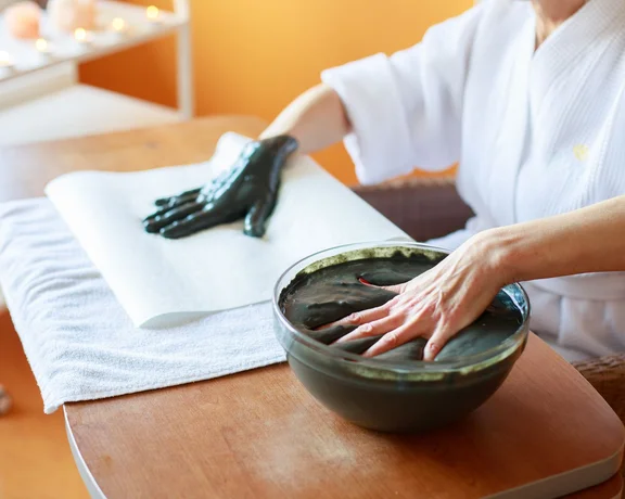 Woman’s hands during a spa treatment for tired skin, one hand resting with a mud mask applied, the other hand beginning to apply the mask.