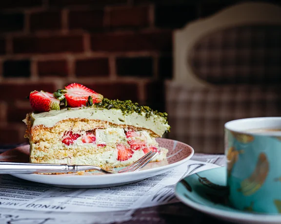 "A slice of cake topped with fresh strawberries on a small plate in a café, accompanied by a cup of tea.