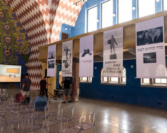 Interior of an exhibition hall with banners detailing Jewish art and freedom struggles, featuring a video screen and rows of empty clear chairs.