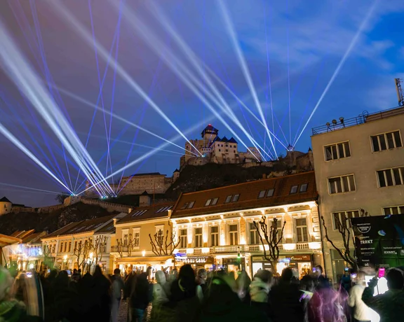 Spectacular night view of Trenčín Castle illuminated by white and blue laser beams during a public show, with a crowd watching below.