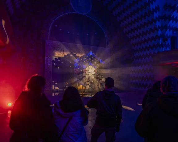 People watching an illuminated geometric light art installation inside a dark, richly decorated hall, featuring red and blue uplighting.