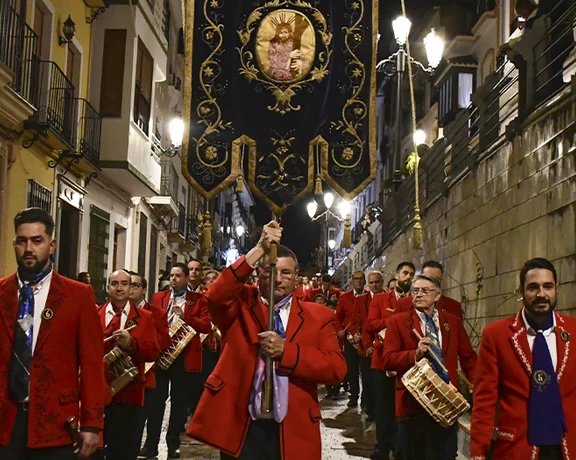 Marching band in red jackets parading down a narrow, well-lit street at night.