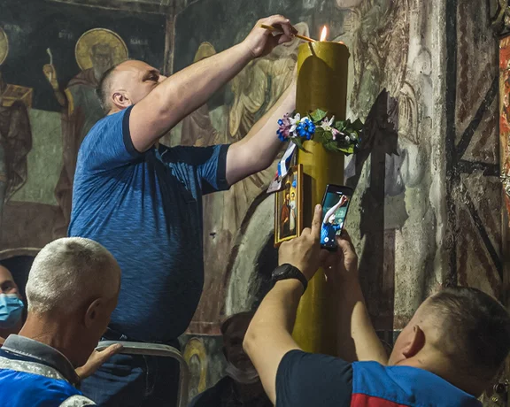 Men lighting a large decorated candle inside an ornate church with religious paintings.