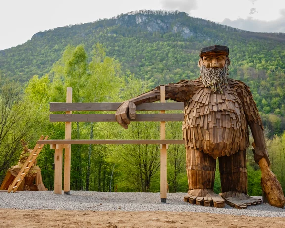 Large wooden troll sculpture sitting on a bench in a green mountainous park.
