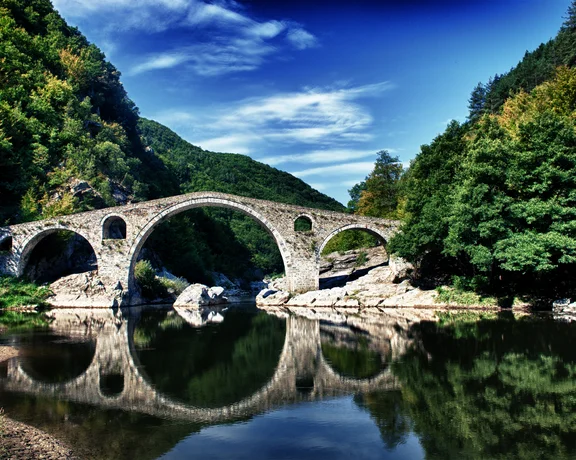 Stone arch bridge over a calm river, surrounded by green trees and hills under a blue sky.