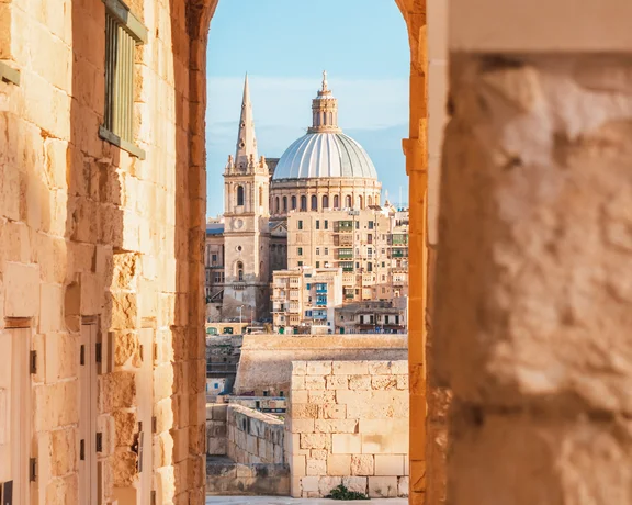 Stone archway framing a distant cityscape with a domed church and spire at sunset.