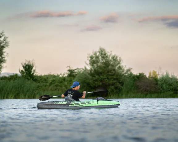 Person kayaking on a calm river at sunset, surrounded by green trees and reeds.