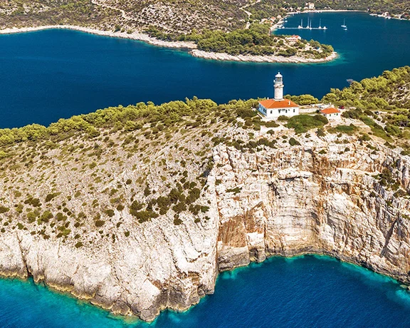Rocky coastline with a white lighthouse surrounded by blue sea and green trees.