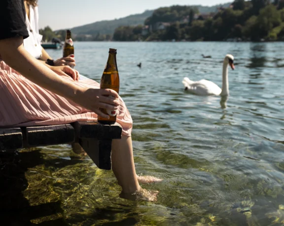 Person sitting on a dock with feet in water, holding a beer, near a swan on a lake.